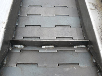 A chain plate conveyor belt with flat surface on the ground. Side plates and transverse baffle being welded onto the belt.